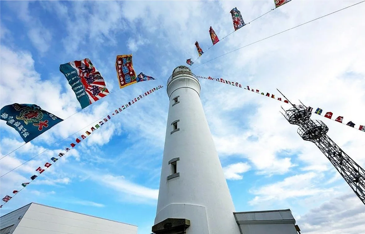 青い空と白い雲の下、白い灯台がそびえ立ち、周囲には日本の旗やカラフルな小旗が飾られ、祝祭的な雰囲気を醸し出しています。