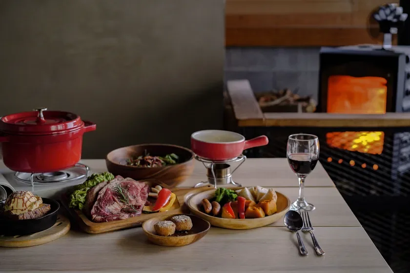 暖炉の炎が温かい雰囲気の店内で、ローストビーフやフォンデュ、鍋料理、ワインなどが並んだ豪華なディナーのテーブル風景です。特別な日の食事にぴったりな、彩り豊かな料理が食欲をそそります。