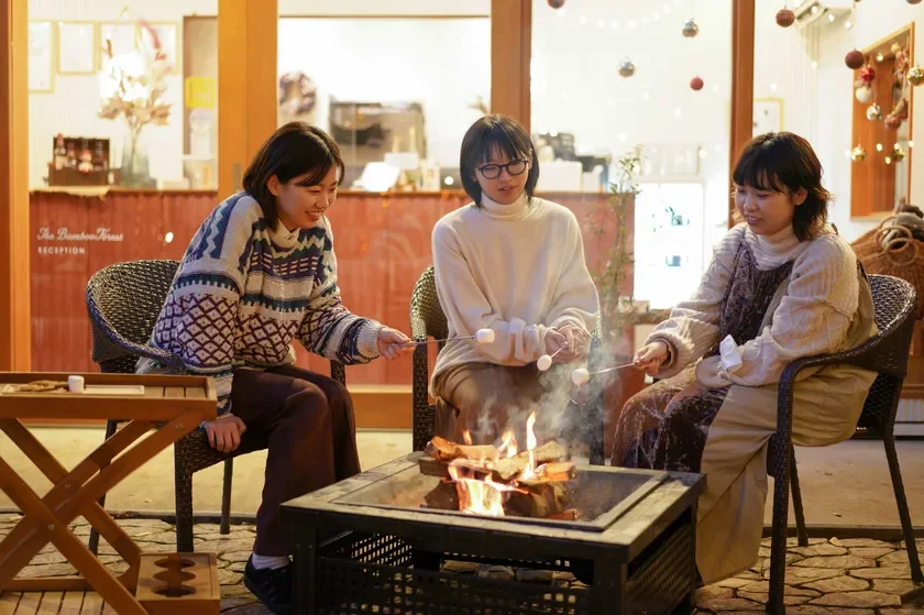 3人の女性が屋外で焚き火を囲み、マシュマロを焼いて楽しんでいる様子です。冬の夜、暖かな火を囲んで友人たちと過ごすリラックスした時間が伝わってきます。