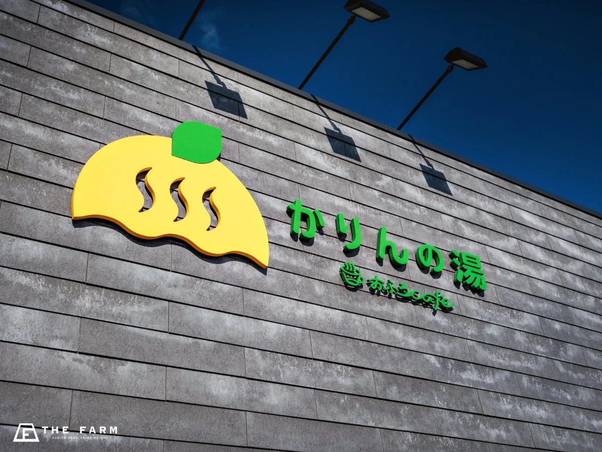 青空の下、グレーの壁の建物に「かりんの湯」と「あかももcafe」の看板が掲げられている。黄色いロゴマークが特徴的な温浴施設とカフェの外観で、千葉県香取市のアグリズムパーク内にある。