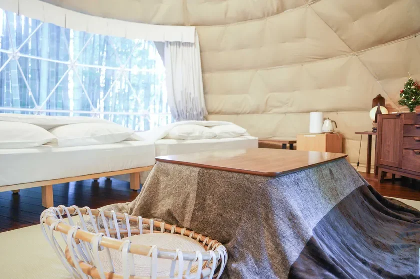 ドーム型の宿泊施設の一室で、白いベッドが2台とこたつが配置されています。大きな窓からは自然光が差し込み、奥には小さなクリスマスツリーも見え、居心地の良い空間が広がっています。
