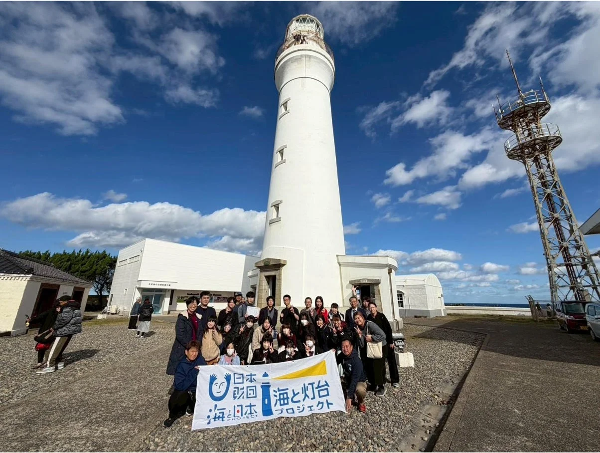 「日本財団 海と灯台プロジェクト」の旗を持つ人々が、晴天の下、白い灯台の前で集合写真を撮っています。