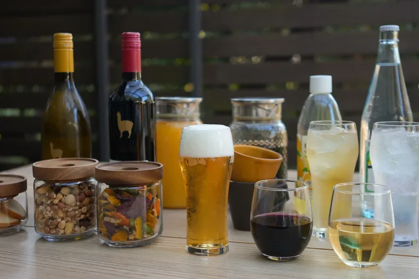 様々な種類の飲み物(ビール、ワイン、ジュース、水など)と、ナッツやドライフルーツなどのおつまみがテーブルに並べられています。カジュアルな雰囲気で、友人との集まりやカフェでのひとときを思わせる光景です。