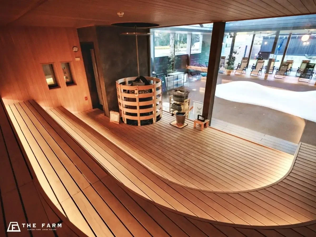 木製のベンチと大型サウナヒーターが特徴のモダンなサウナルームです。大きな窓からは屋外のリラックスエリアが見え、暖かみのある照明が心地よい空間を演出しています。