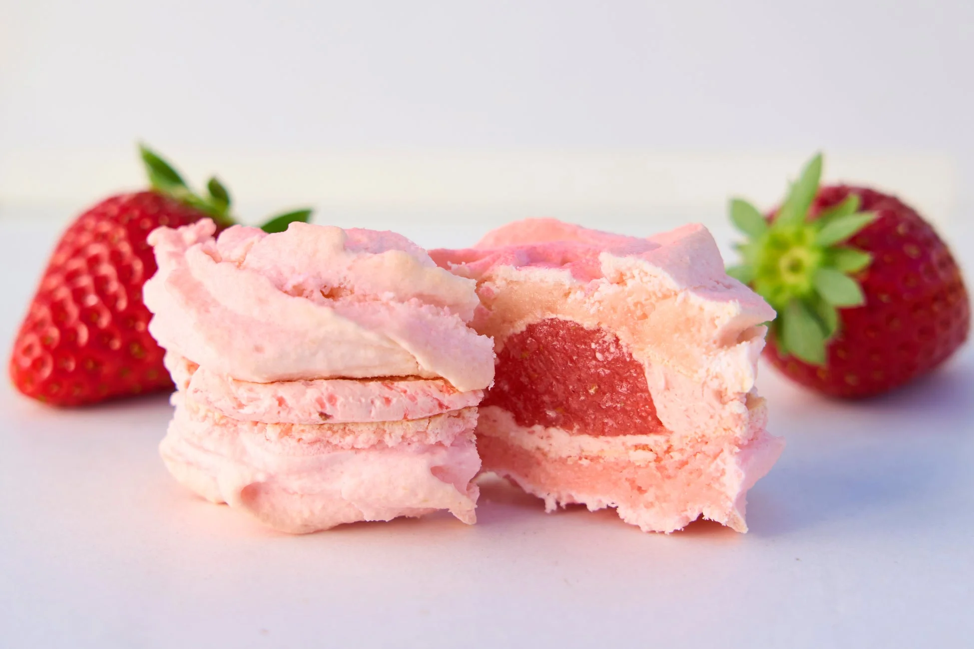 白い背景に、ピンク色のメレンゲ菓子と新鮮ないちごが並んでいます。メレンゲ菓子は半分にカットされ、中に赤いフィリングが見えており、甘く美味しそうな雰囲気を醸し出しています。