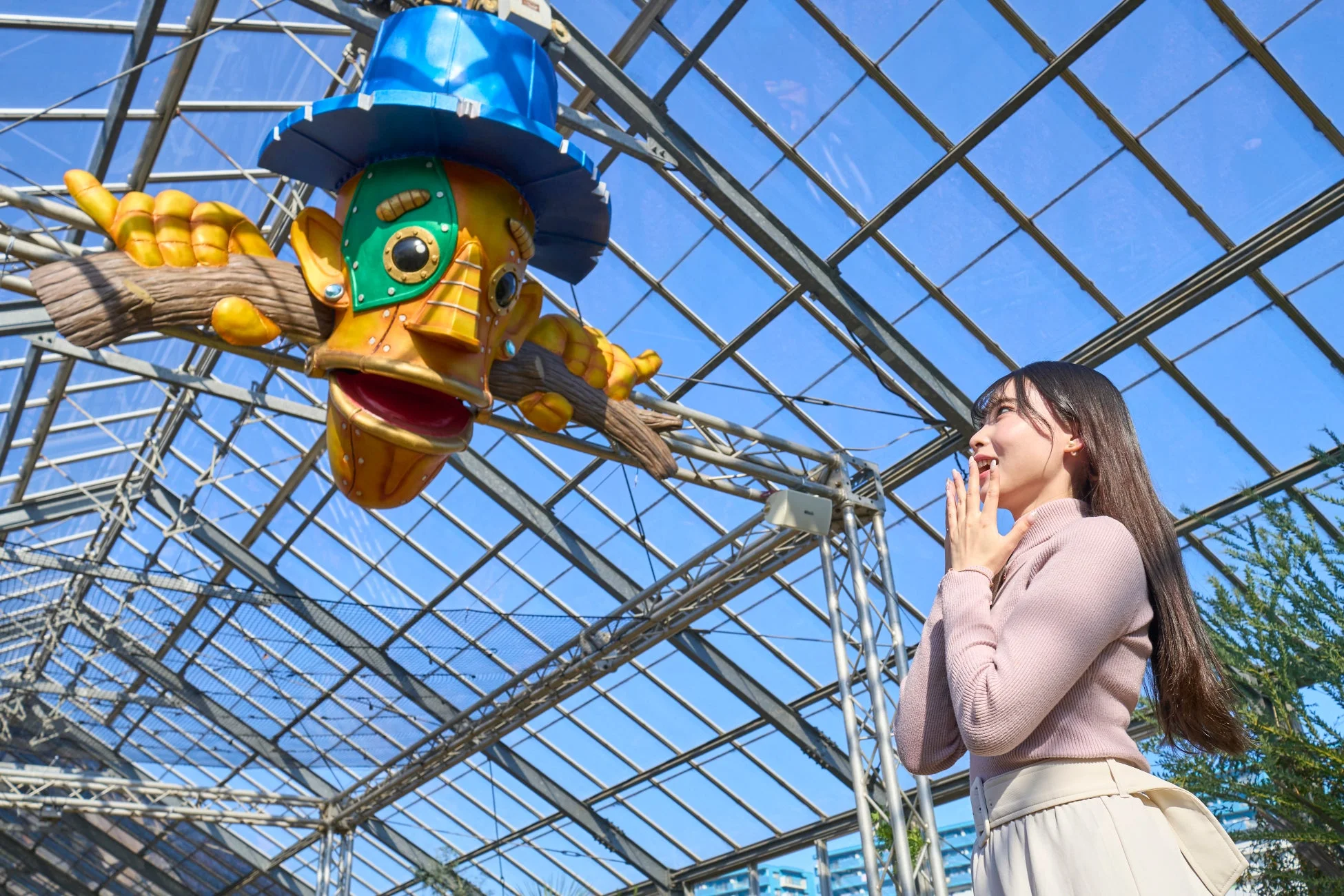 ガラス屋根の明るい空間で、若い女性が頭上に吊るされたカラフルで大きな魚のようなキャラクターを見上げています。女性は驚きと喜びの表情を浮かべており、開放的な施設での楽しいひとときを捉えた一枚です。