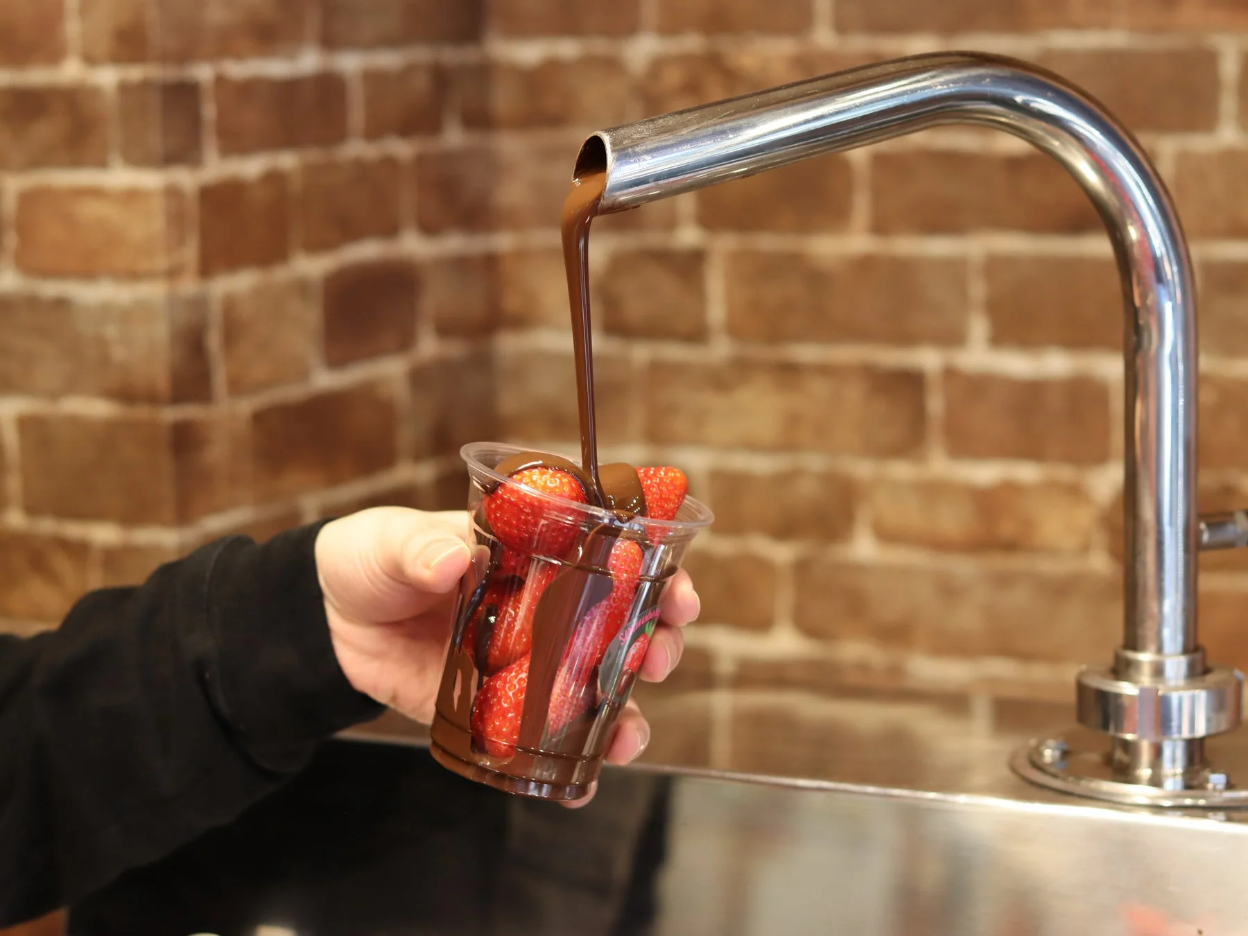 透明なカップに入った新鮮なイチゴに、とろけるチョコレートが上からたっぷりと注がれている瞬間を捉えた画像です。背景にはレンガの壁が見え、甘く美味しそうなデザートが作られている様子が伝わってきます。