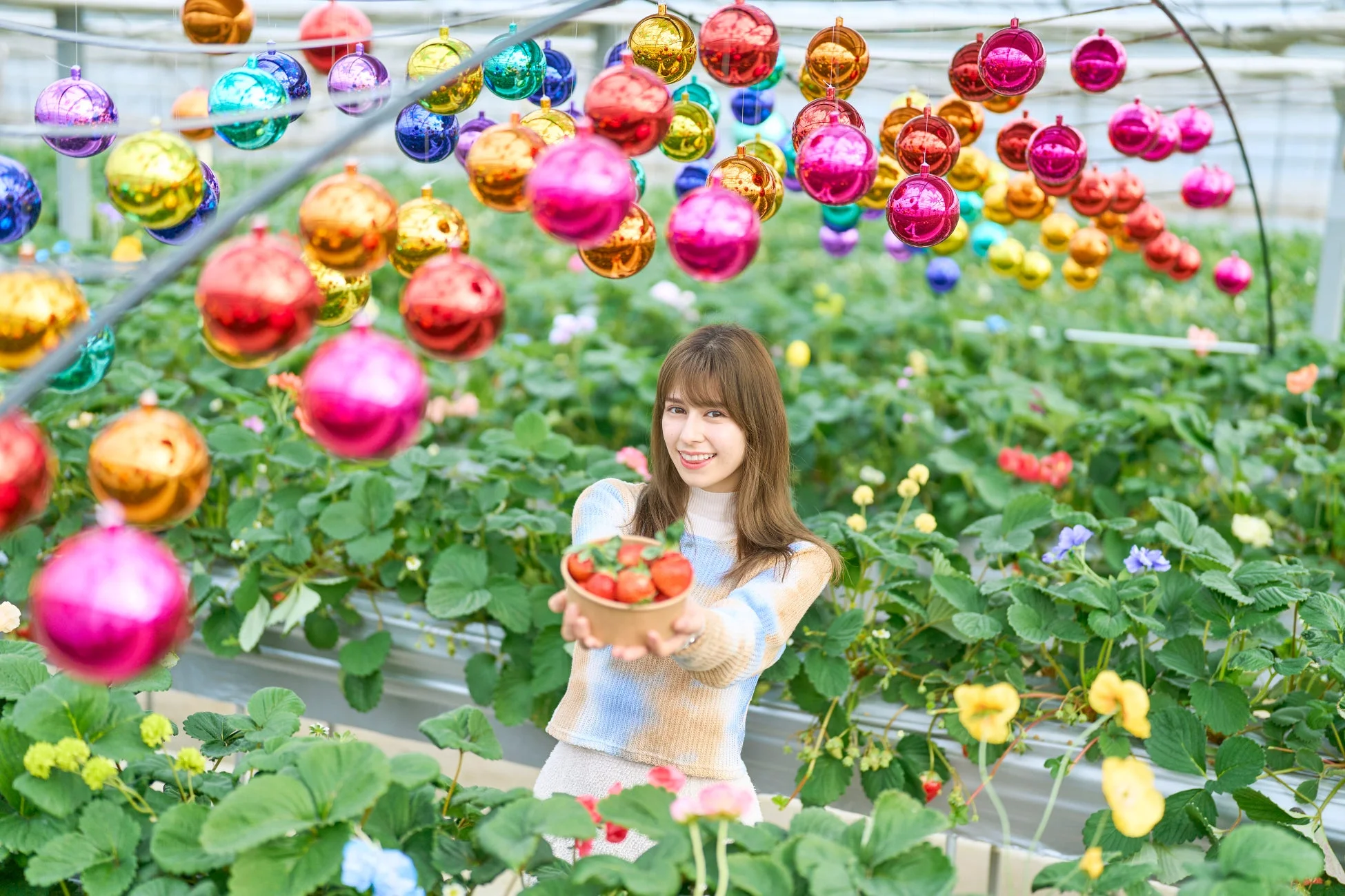 イチゴ農園のような場所で、笑顔の女性が摘みたてのイチゴが入ったボウルを差し出している。頭上にはカラフルなオーナメントが吊るされ、華やかな雰囲気を醸し出している。