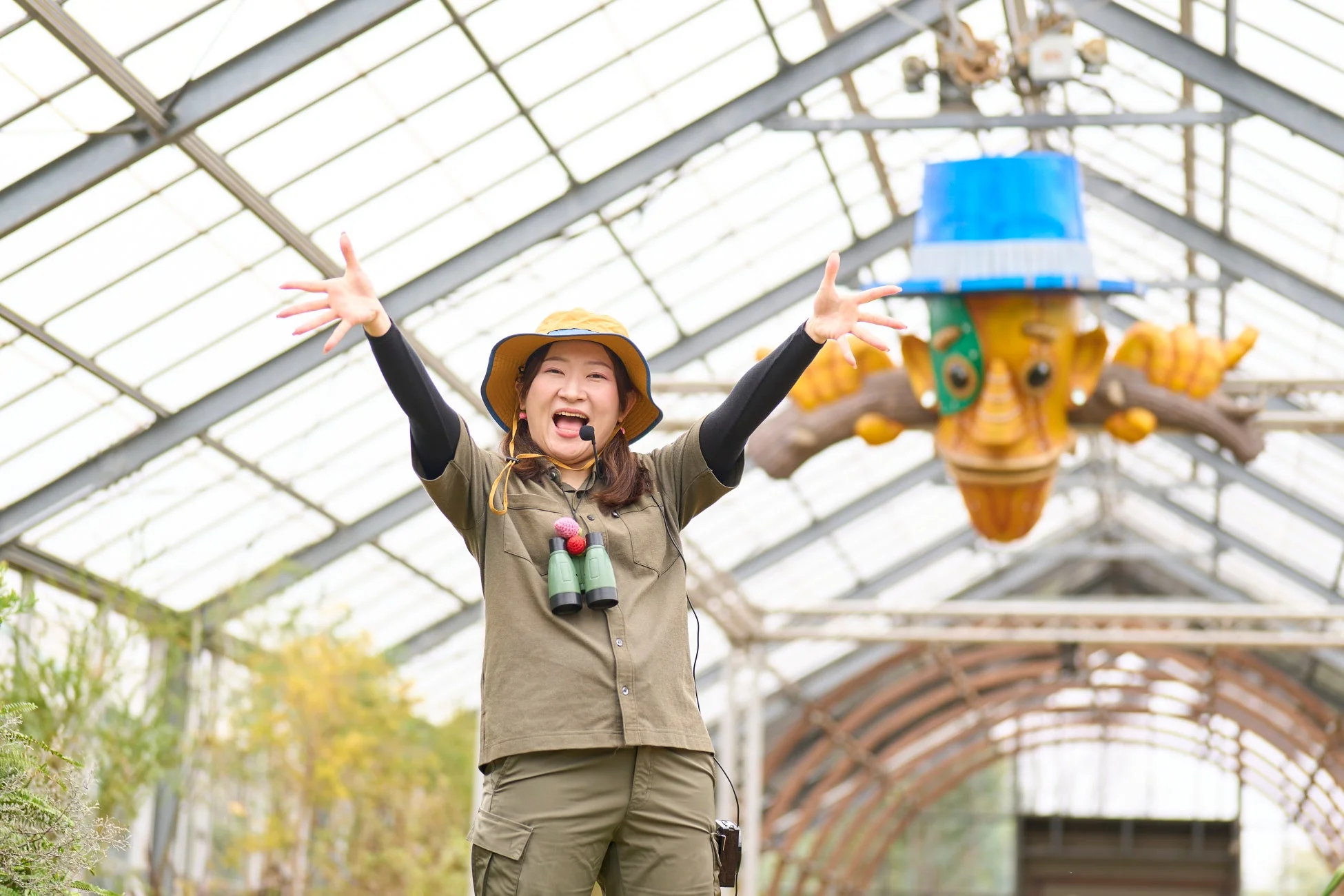 サファリ服を着た女性が温室のような場所で両腕を広げ、満面の笑みで何かを説明している。双眼鏡とマイクを身につけており、楽しそうなイベントの様子を伝えているようだ。