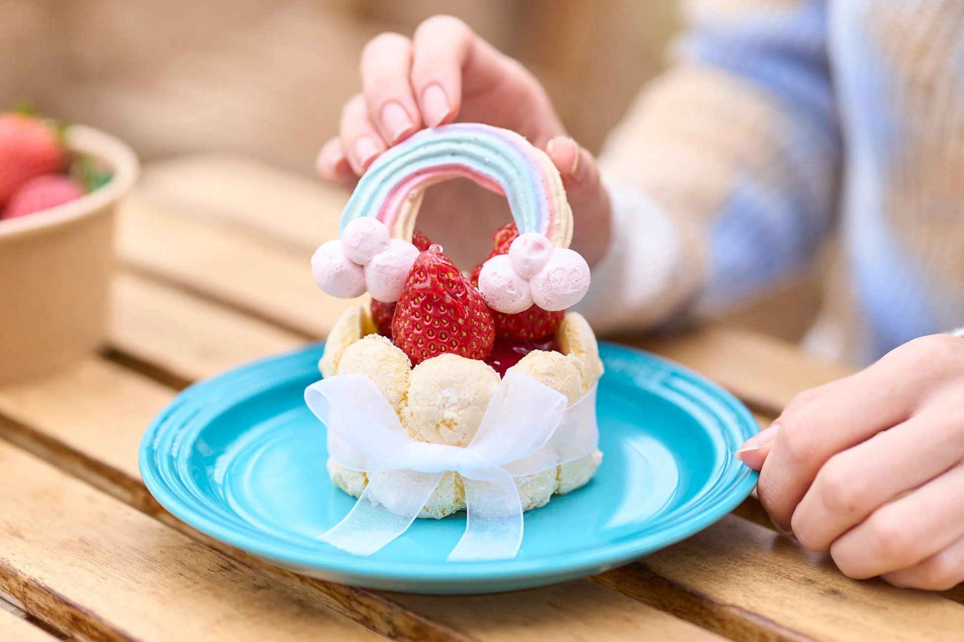 木のテーブルに置かれたターコイズ色の皿の上で、白いリボンで飾られた可愛らしいイチゴのシャルロットケーキに、手で虹色のメレンゲを添えている様子。背景にはイチゴのボウルも見えます。