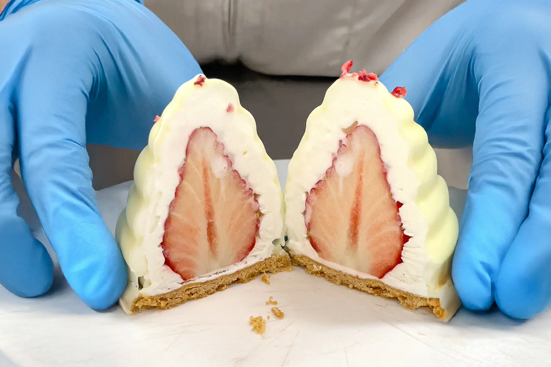 青い手袋をはめた手が、丸ごといちごが中に入った白いクリームのデザートを半分に割って見せています。サクサクのタルト生地の上に、たっぷりのクリームとみずみずしいいちごが乗った美しい断面が特徴的なスイーツです。