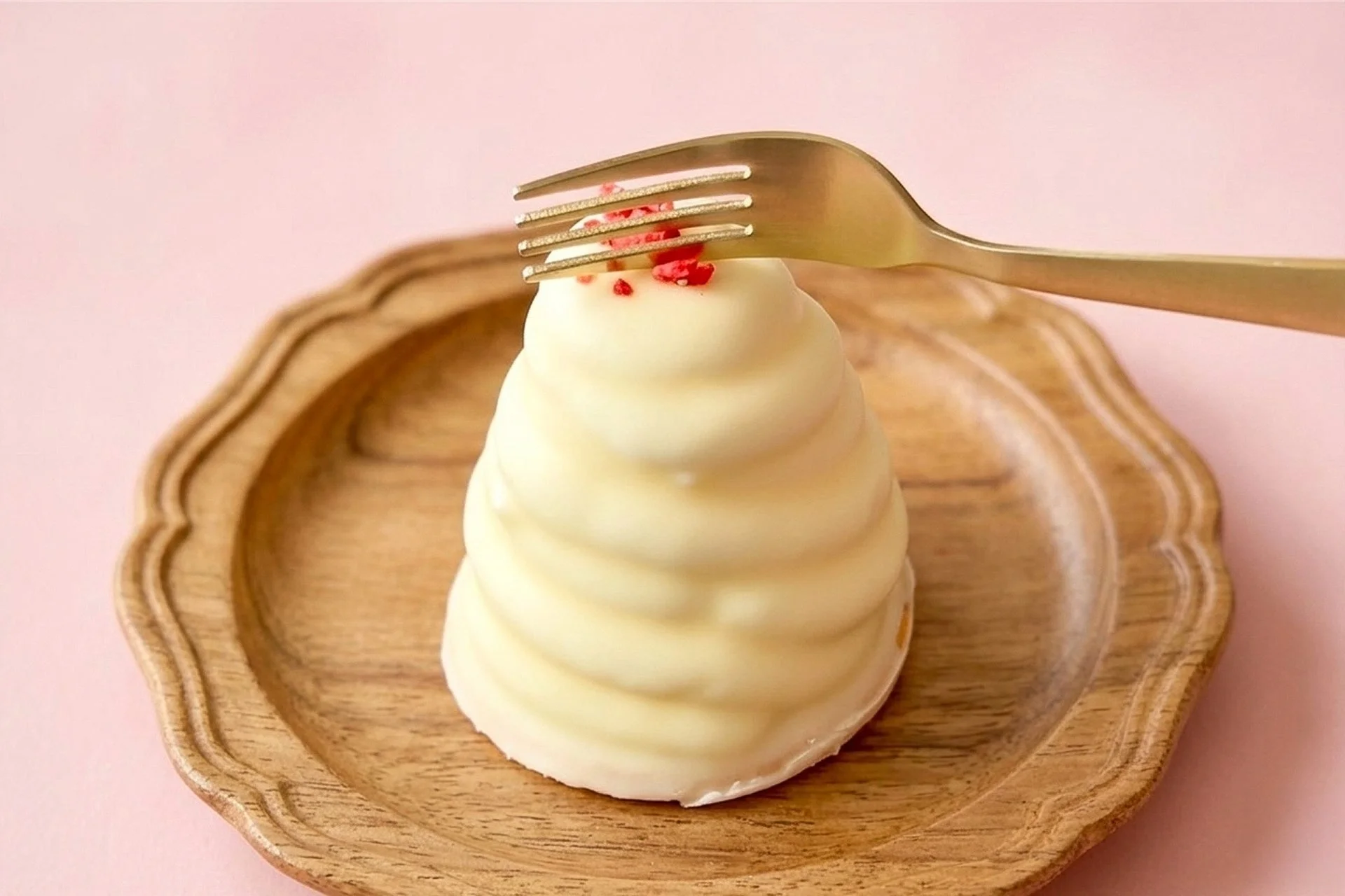 白い渦巻き模様が特徴的な可愛らしいデザートが木製の皿に乗せられ、その上には金色のフォークが置かれています。淡いピンクの背景が、全体の甘く優しい雰囲気を引き立てています。