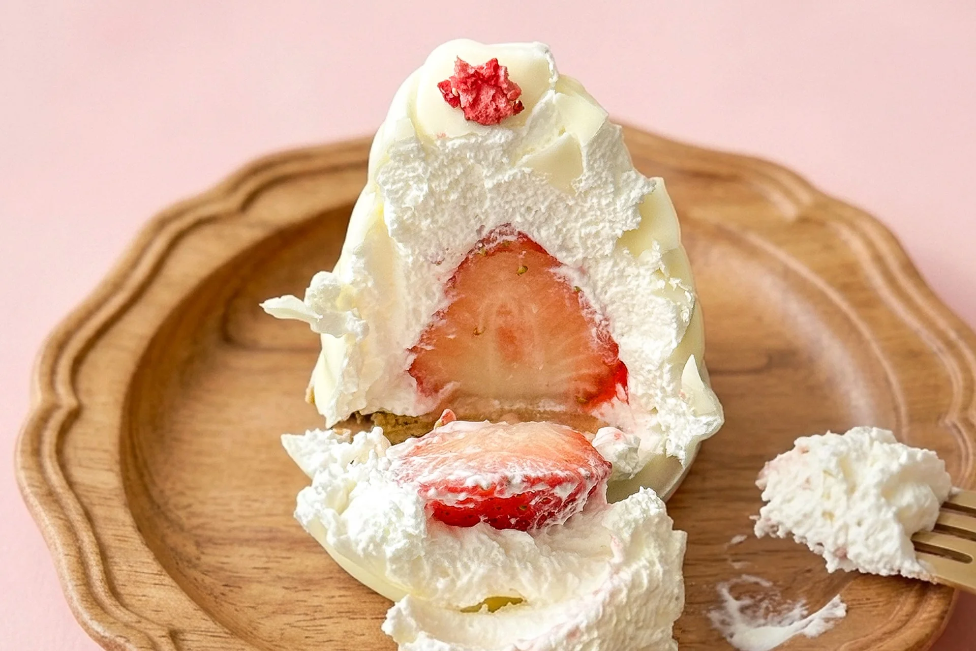ピンクの背景の木製プレートに、生クリームと白いシェルで包まれたいちごのデザートが置かれています。崩れた断面から瑞々しいいちごが見え、甘く美味しそうな雰囲気が伝わってきます。