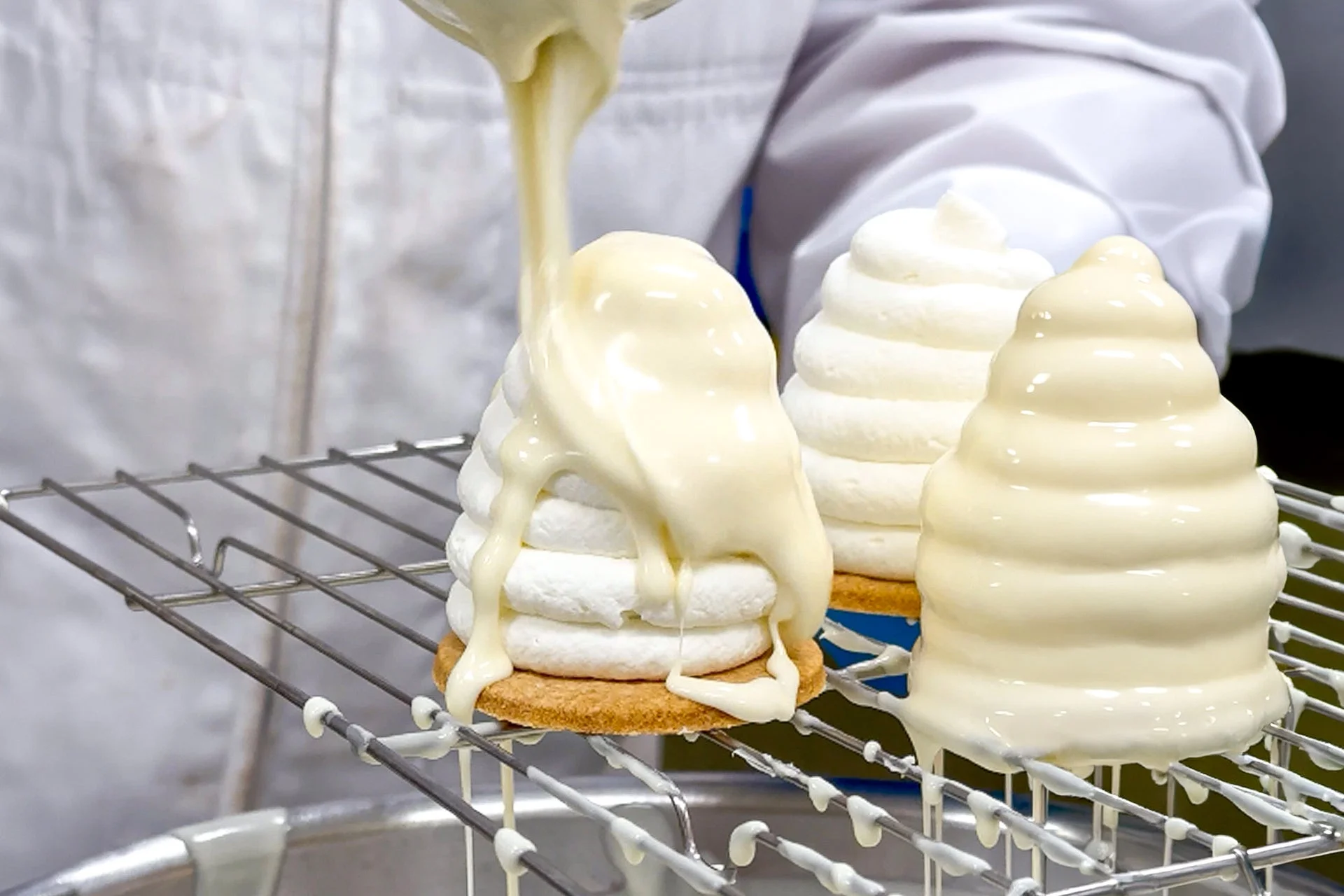 白い制服を着た人が、冷却網に置かれたビスケットベースの円錐形デザートに白いソースをかけている様子です。他にも同様のデザートが並んでおり、菓子作りの最終工程を示しています。