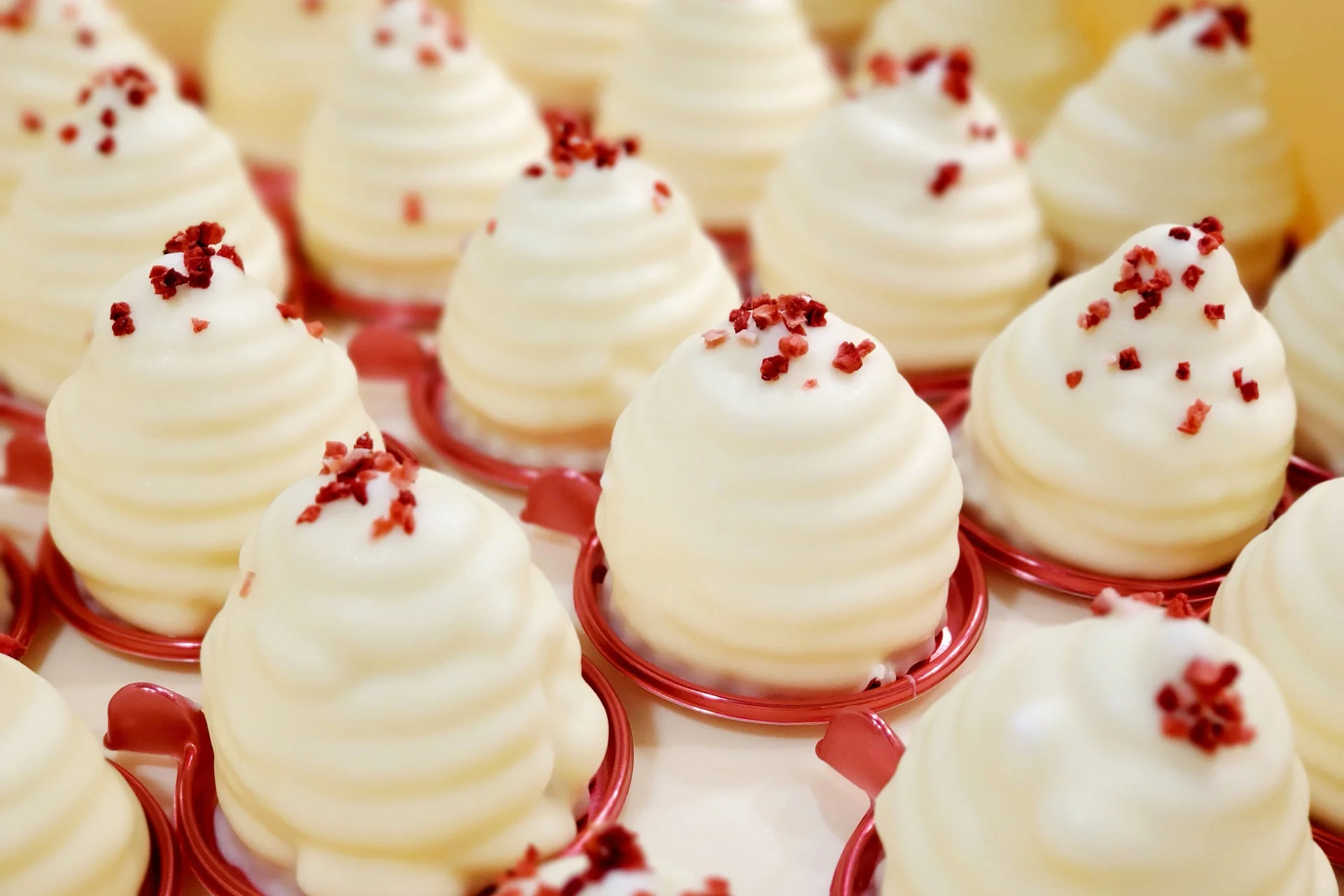 白い螺旋状の可愛らしいデザートが多数並んでいます。赤い粒で飾られた一つ一つのお菓子は、赤い土台に乗っており、お祝いやギフトに最適な甘い雰囲気を作り出しています。