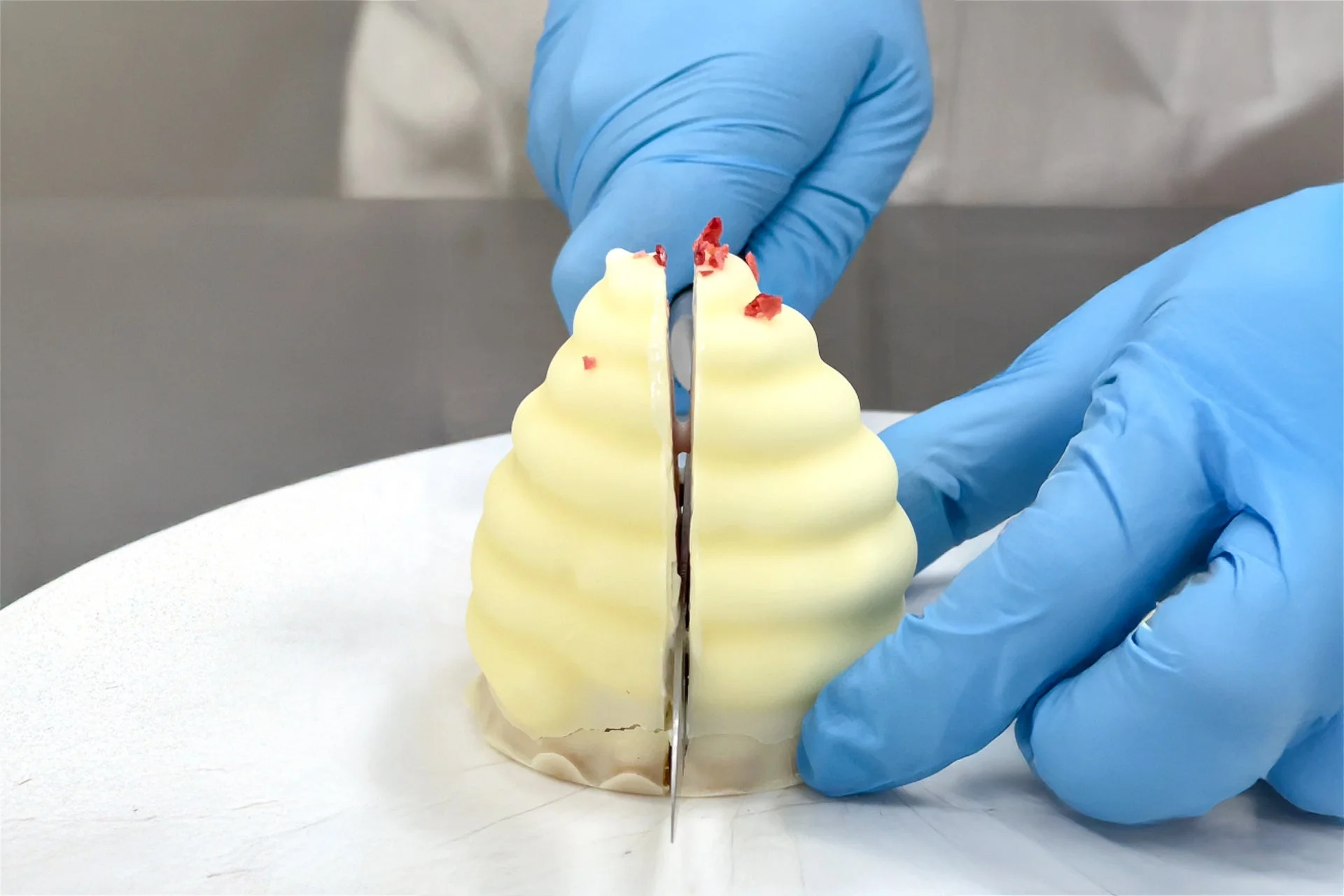 青い手袋をはめた手が、白いクリーム状のデザートをナイフで中央からカットしている様子。デザートは渦巻き状で、上部には赤い小さなトッピングが見えます。プロフェッショナルな作業風景です。