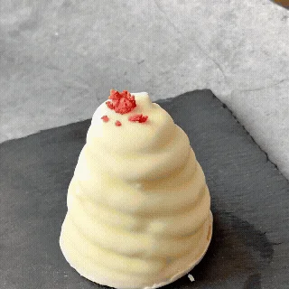 白いクリームで覆われた円錐形のデザートが、黒いプレートに置かれています。上部には赤い小さなトッピングが散りばめられ、螺旋状の模様が特徴的です。